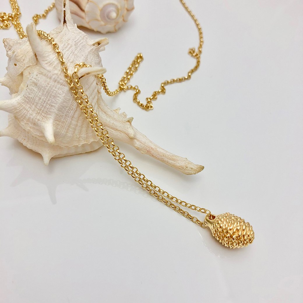 A delicate gold chain necklace features a textured gold pendant, elegantly draped over a large, spiky white seashell. In the background on a plain white surface, another seashell and part of the chain can be seen beside a baby pine cone. This beautiful piece of jewellery, the Pine Cone Long Necklace by Jennifer Kinnear, captures natural elegance effortlessly.