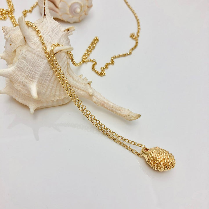 A delicate gold chain necklace features a textured gold pendant, elegantly draped over a large, spiky white seashell. In the background on a plain white surface, another seashell and part of the chain can be seen beside a baby pine cone. This beautiful piece of jewellery, the Pine Cone Long Necklace by Jennifer Kinnear, captures natural elegance effortlessly.