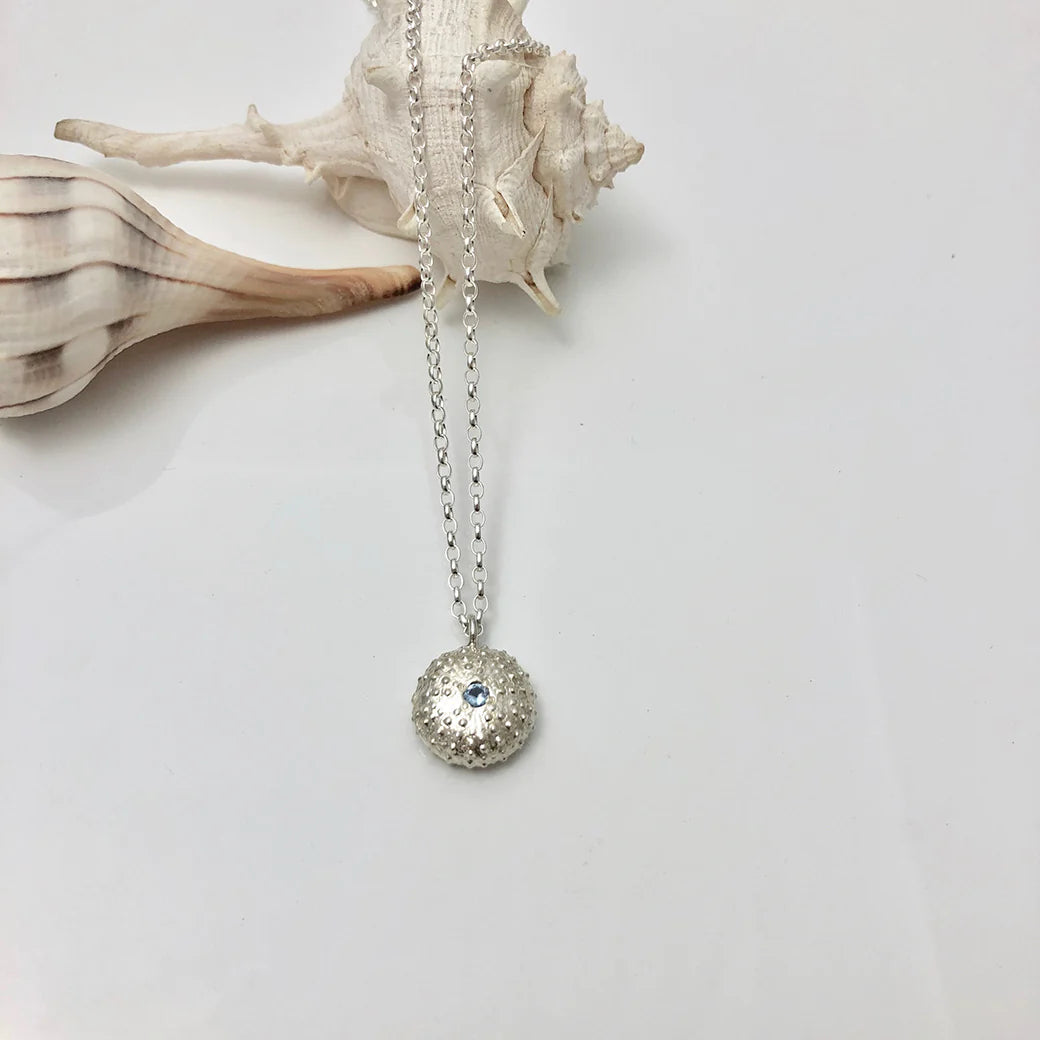 The Jennifer Kinnear Sea Urchin Necklace with Stone (silver) features a textured, round pendant with a small blue stone at the center, shown on a white surface beside two seashells.