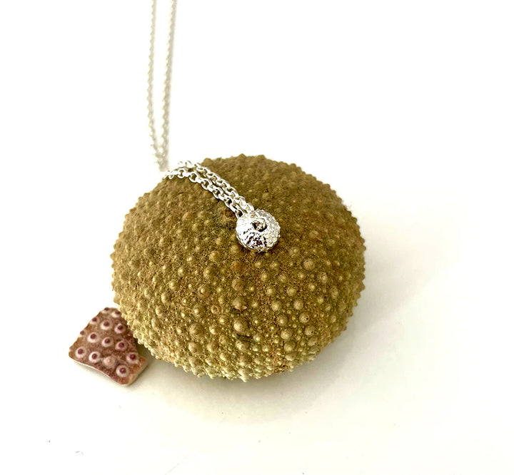 The Jennifer Kinnear Sea Urchin Necklace Tiny (Silver or Gold) features a delicate, textured pendant displayed on a spiky sea urchin shell next to pink coral, all set against a white background.