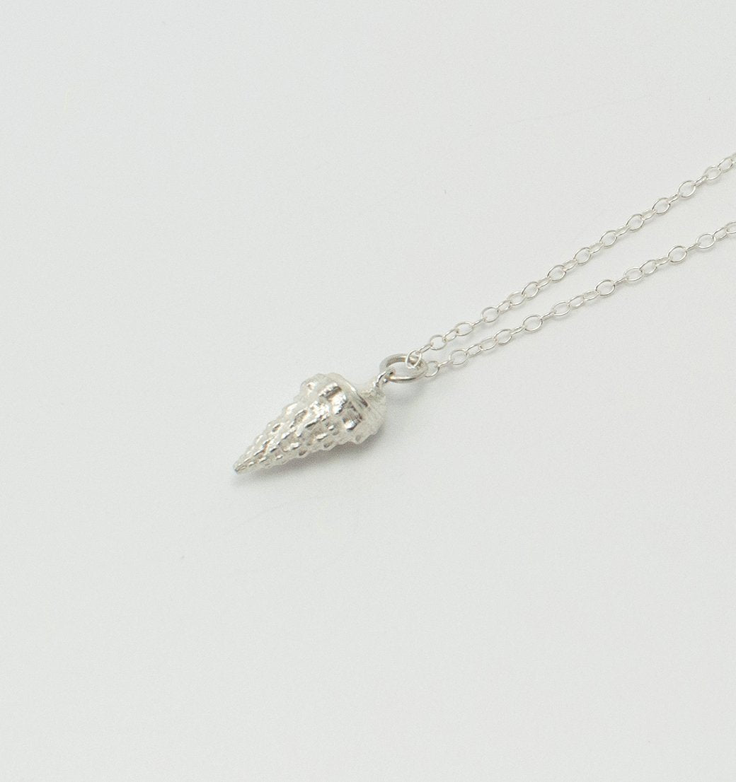 A delicate **Whelk Shell Necklace** by **Jennifer Kinnear** features a small pendant shaped like an ice cream cone, reminiscent of summer days in the Balearic Islands. The intricate details mimic a textured cone with a scoop of ice cream on top. The fine, simple chain perfectly complements the whimsical pendant design.