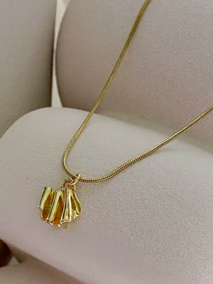 The Little Joy Necklace by Inner island features a thin gold chain with a pendant of three textured, elongated gold shapes, displayed on a soft beige jewelry stand—an elegant choice for everyday wear.