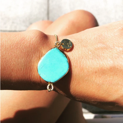 A Juno bracelet by Aria V, featuring a large turquoise stone centerpiece and a small round gold charm on an adjustable gold plated brass chain, is worn on a person's wrist. The background is blurred, focusing on the jewelry and the wrist.