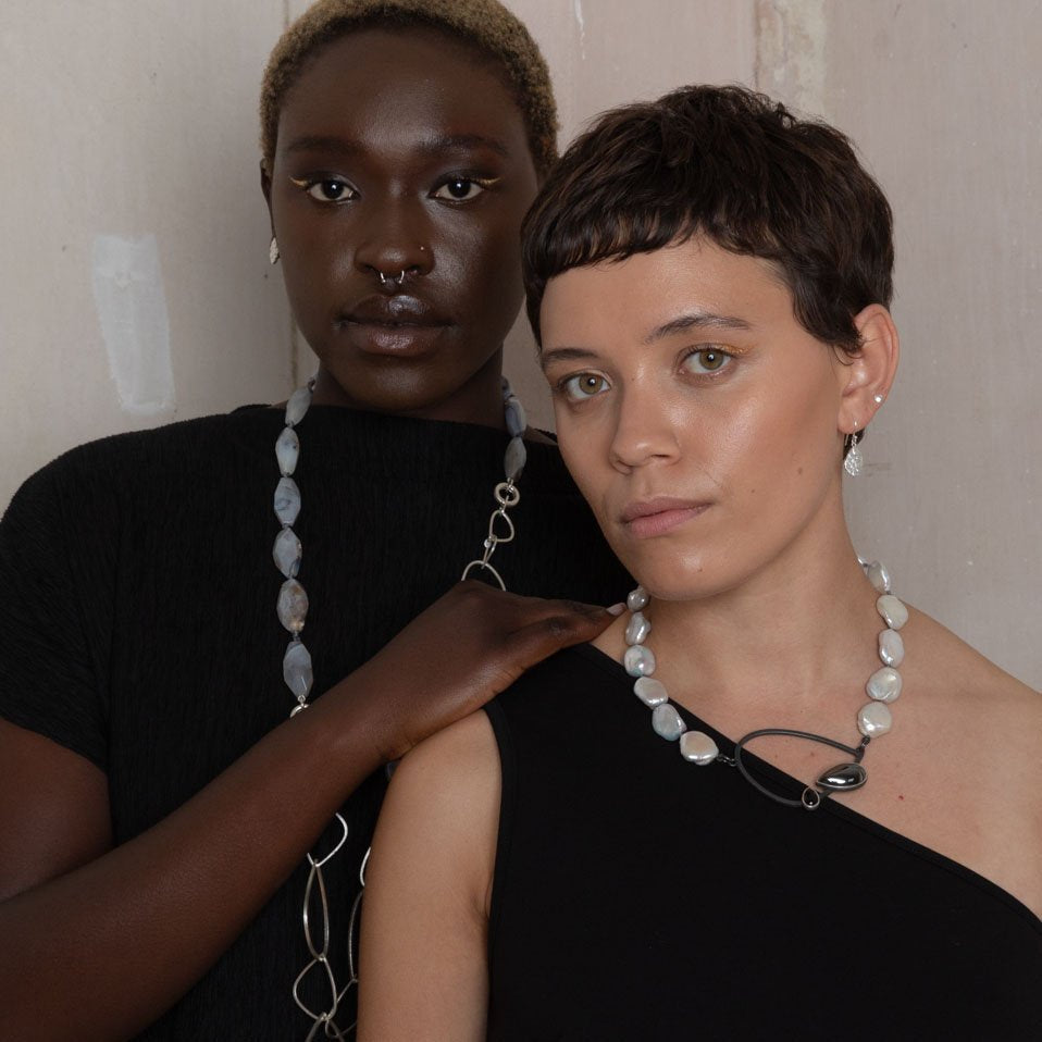 Two individuals are posing against a neutral background. They both wear statement necklaces with large, white beads and minimalistic clothing. The person on the left has their hand gently resting on the shoulder of the person on the right, who is also wearing elegant Kimana Drop Earrings in Frosted Silver from Leoma Drew.