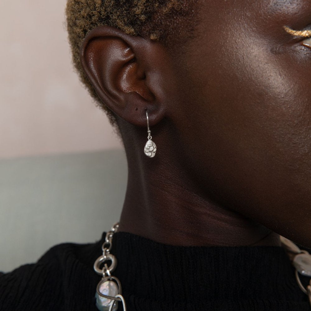 Close-up of a person wearing **Leoma Drew Kimana Drop Earrings in Frosted Silver** shaped like a small teardrop with intricate detailing. The person's skin is dark, and they have short, curly hair. They are also wearing a chunky silver necklace. The background is softly blurred, highlighting the elegant drop earrings.
