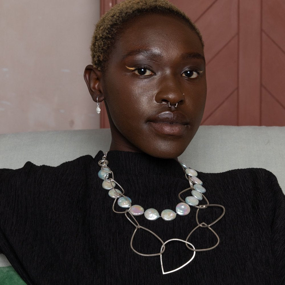 A person with short blonde hair and dark skin is seated, wearing a statement necklace made of large pearls and metal shapes. They have a nose ring, Kimana Drop Earrings in Frosted Silver by Leoma Drew, and are dressed in a black top. The background includes a textured wall and a wooden door.