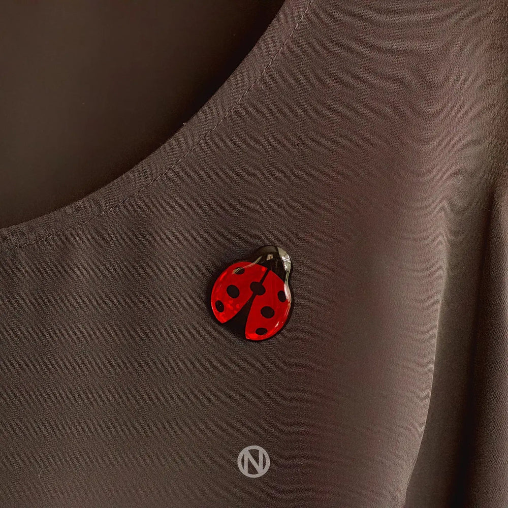 A small, hand-painted Ladybird Pin Brooch with black spots is pinned on a dark-colored fabric, possibly a shirt. The whimsical touch of the ladybug contrasts beautifully with the plain background. A logo featuring an "N" within a circle is visible at the bottom of the image, adding to its distinct charm from Naoi.