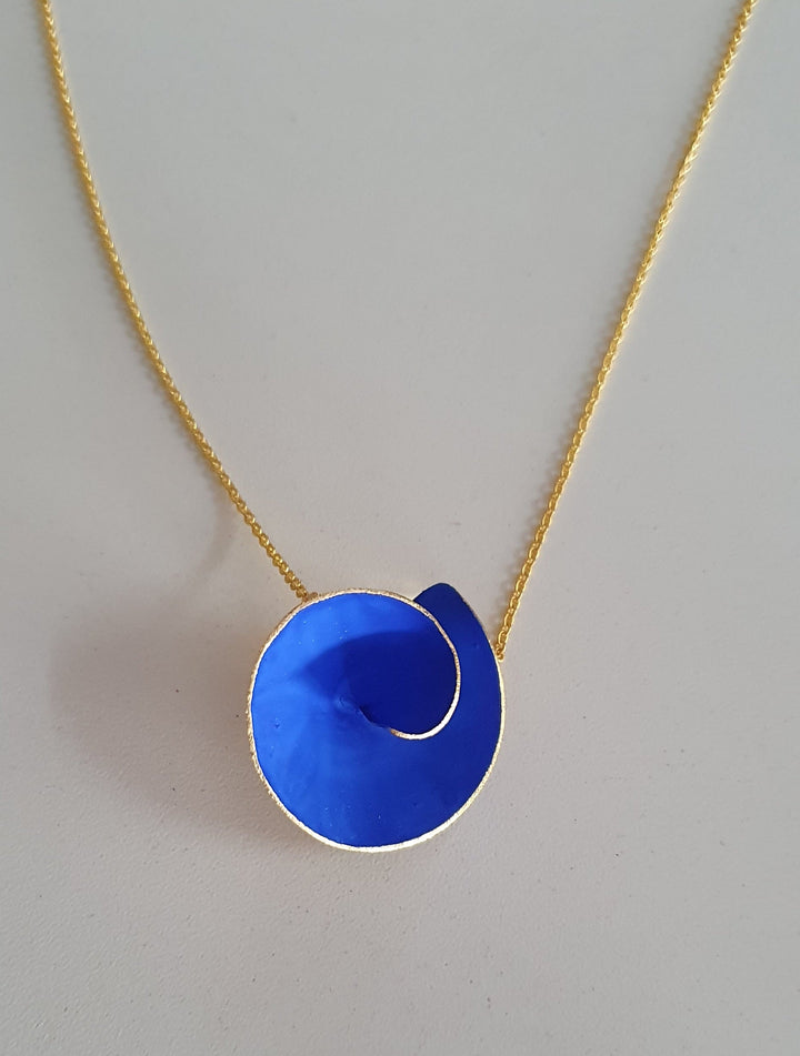 Close-up of a hand made necklace with a gold plated silver chain and a circular, cobalt blue pendant that is gold-edged on the outside, resting on a light gray surface. This Large Matt Swirl Pendant by Georgia Charal adds an exquisite touch to any outfit.