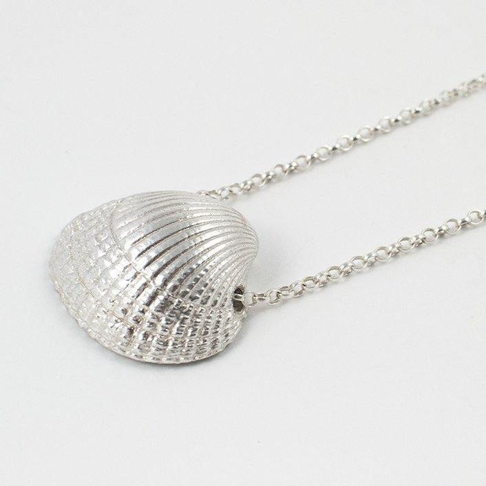 The Jennifer Kinnear Cockle Shell Necklace Large, offered in both silver and gold, features a detailed, textured seashell pendant that hangs elegantly on a fine silver chain. The plain white background highlights the intricate charm of this beautifully crafted piece.