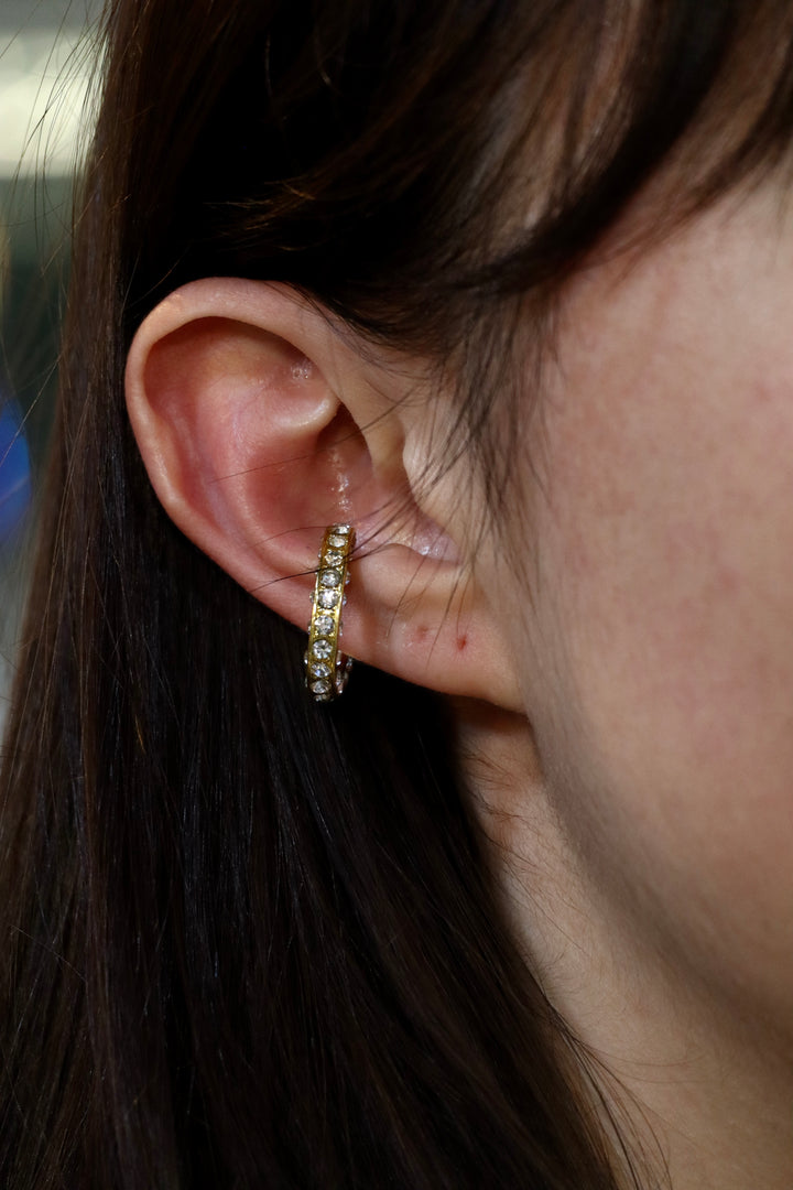 Close-up of a person's ear with straight brown hair, wearing a gold hoop earring and the Zubeldia Oval Crystal Gold Plated Ear Cuff. The background is blurred.