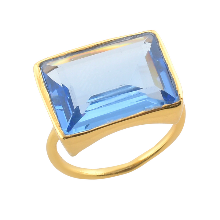 A 22 carat gold ring featuring a large rectangular blue gemstone set in a bezel setting. The faceted cut of the gemstone gives it a brilliant reflective surface, making it an eye-catching statement piece. The ring band is simple and smooth. Perfect as a cocktail ring, the **Lenny Ring** by **Shyla** is sure to impress.