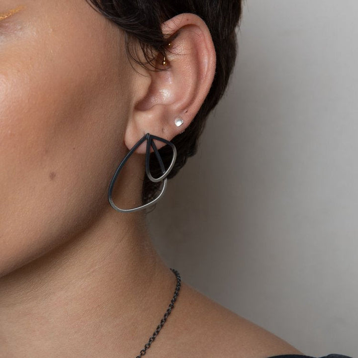 Close-up of a person with short hair wearing Leoma Drew Two Tone Double Wing Studs on their left ear. The earring has a hollow, intertwined design. The person is also wearing a small stud earring above the teardrop one, and part of a necklace chain is visible.