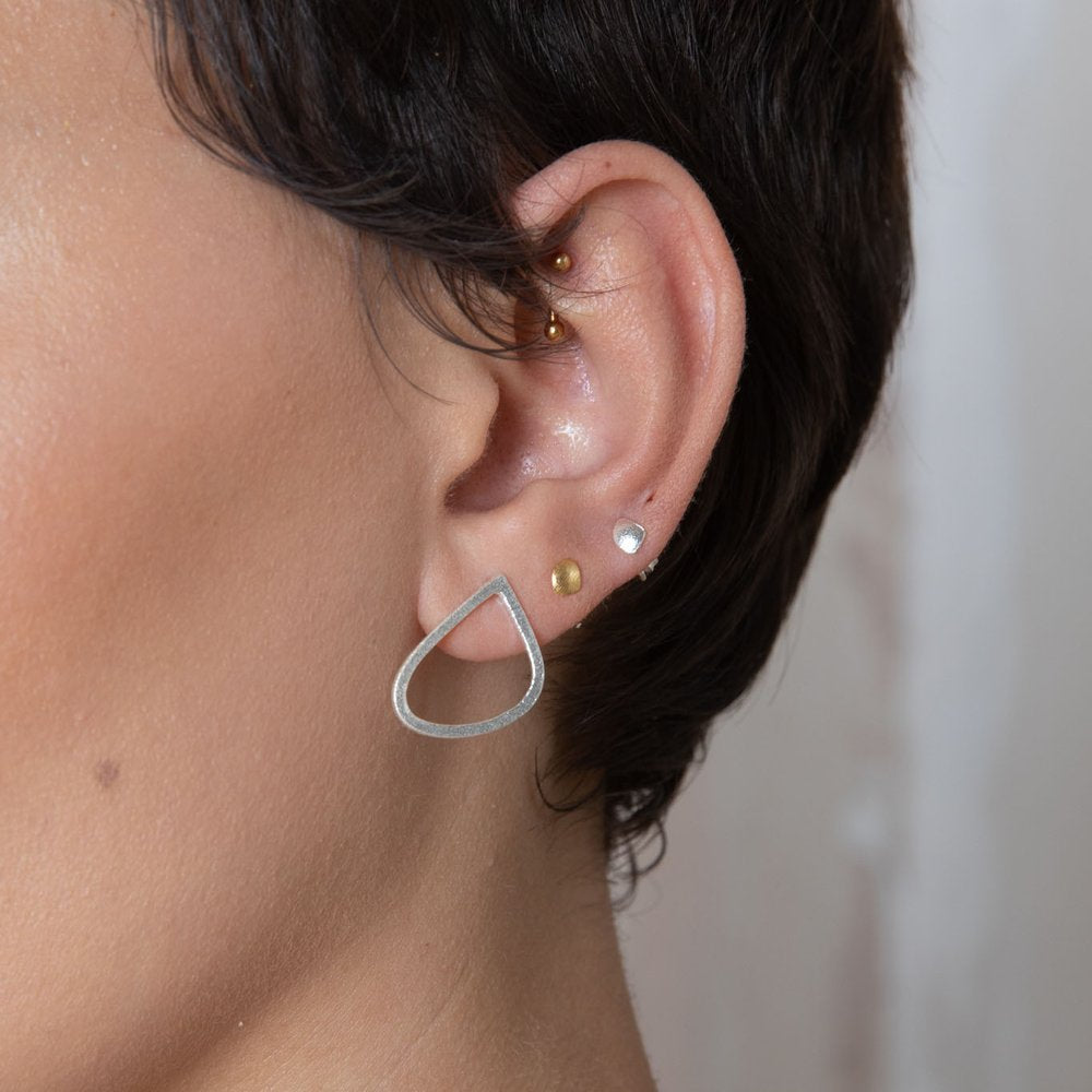 Close-up of a person's ear adorned with multiple piercings. The piercings feature a variety of earrings, including a gold stud, a sterling silver hoop, Leoma Drew Freeform Wing Studs Silver with a frosted finish, and an open teardrop-shaped earring. The person has short, dark hair.