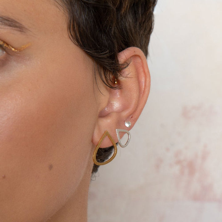 Close-up of a person's ear adorned with multiple earrings. The ear features a small sterling silver stud and two large teardrop-shaped earrings, one gold plated and one silver, layered together. The background is softly lit with a light-colored texture. The sterling silver stud is from the Freeform Wing Studs Gold collection by Leoma Drew.