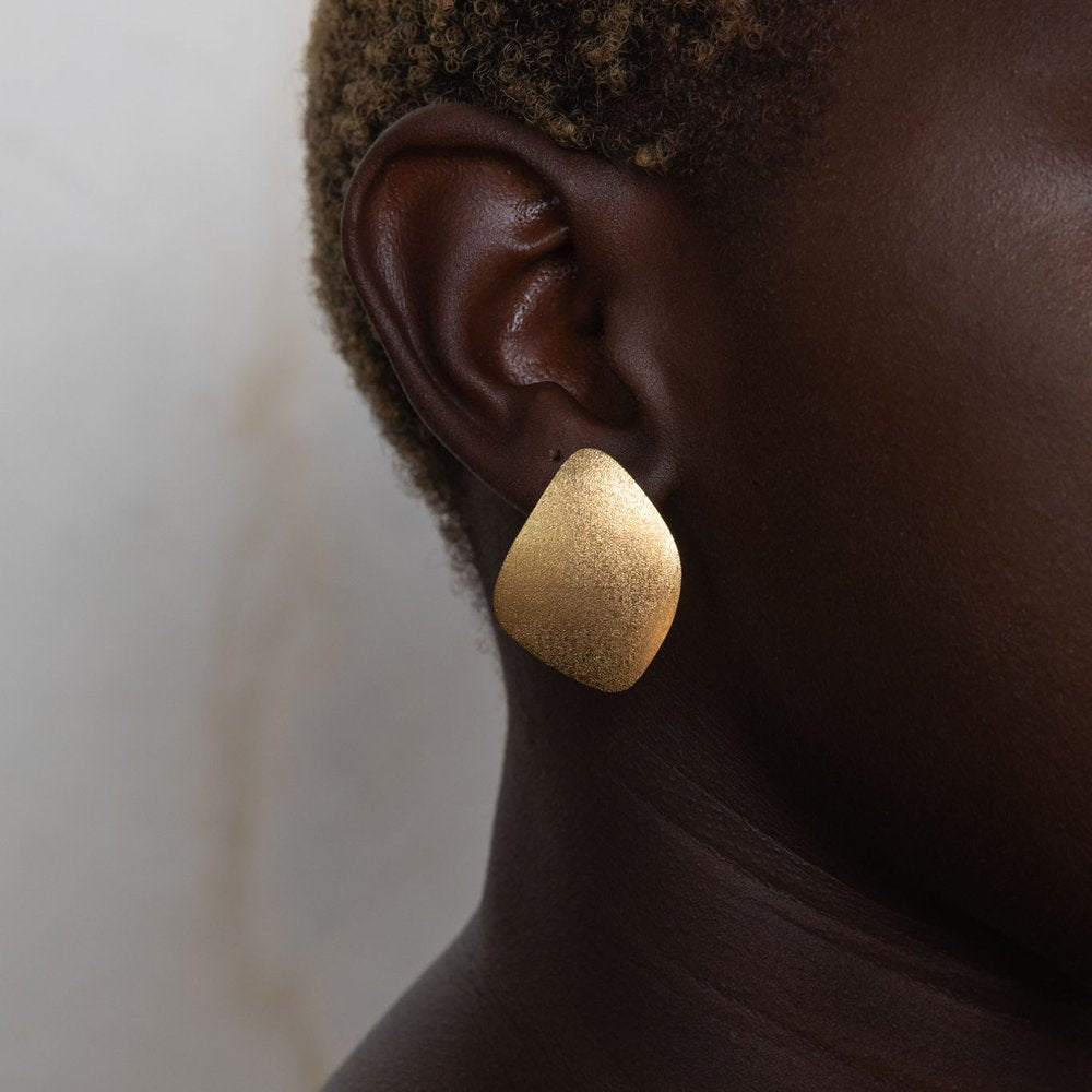 A close-up of a person wearing a single, large, irregularly shaped gold-plated earring. The earring has a frosted finish, and the person's ear and part of the neck are visible against a plain background. The product is called Freeform Wing Studs Gold by Leoma Drew.