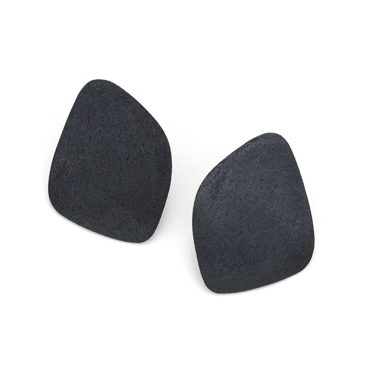 A pair of asymmetrical, black earrings with an organic, irregular shape. Crafted from oxidised sterling silver, each earring features a textured matte finish for a natural, minimalist look. Slightly curved and differing subtly in size and shape from each other, the Freeform Wing Studs Oxidised Silver by Leoma Drew come in four sizes.