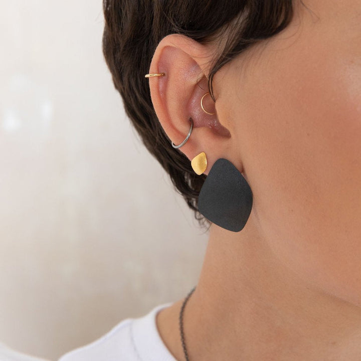 Close-up of a person's ear adorned with multiple earrings, including a small hoop, a triangular gold stud, and Leoma Drew's Freeform Wing Studs Oxidised Silver. The person has short hair and is wearing a white shirt. The background is neutral and blurry.