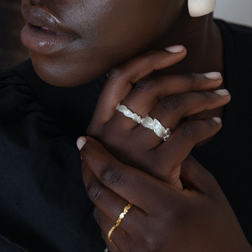 A person with dark skin wearing a black top is shown close-up. They have a cream-colored stone band ring on their middle finger and a thin gold ring with small beads from the Kimana Silver Freeform Layered Wing Ring by Leoma Drew on their pinky. Their hands are gently interlaced near their face.