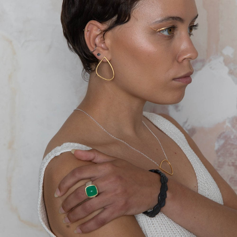 A person with short hair gazes to the side, wearing a white sleeveless top. They accessorize with Gold Wing Stud Earrings from Leoma Drew, a matching necklace, a ring with a green stone, and a black woven bracelet. Their hand rests on their opposite shoulder against a textured background.