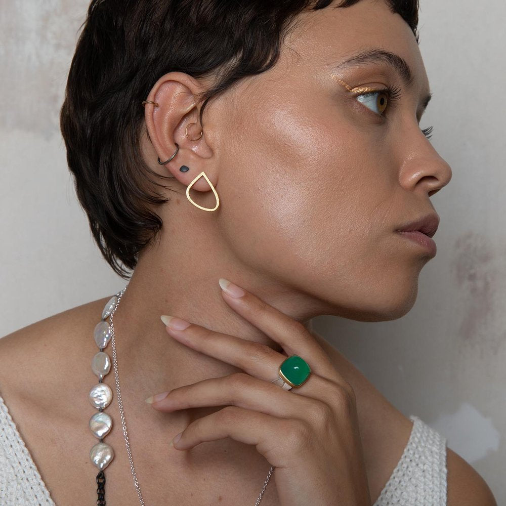 Profile view of a person with short, dark hair looking sideways. They are wearing a textured white tank top, a pearl necklace, and Leoma Drew Gold Wing Stud Earrings. A ring with a large green stone adorns their hand. They have golden eyeshadow and a small black ear piercing.