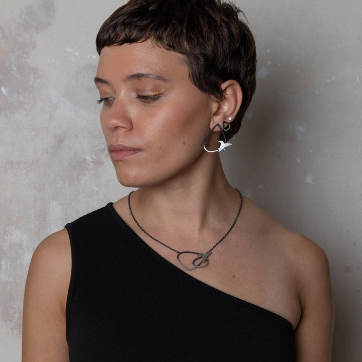 A person with short hair in striking hair colours, wearing a one-shoulder black top, looks to the side. They have Two Tone Silver Wing Studs With Birds by Leoma Drew and a matching necklace in sterling silver, complemented by gold eye makeup. The background is a textured, light-colored wall.