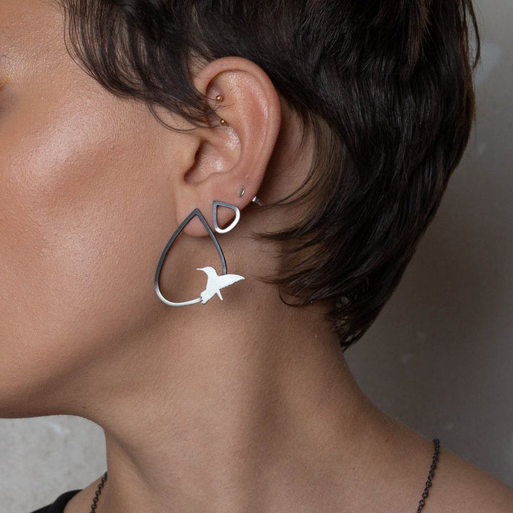 Close-up photo of a person wearing Two Tone Silver Wing Studs With Birds in three sizes by Leoma Drew. The earring is a large, black teardrop shape with a smaller black triangle at the top and features a white, cut-out bird design near the bottom. Crafted from sterling silver, it complements their short hair and multiple ear piercings.