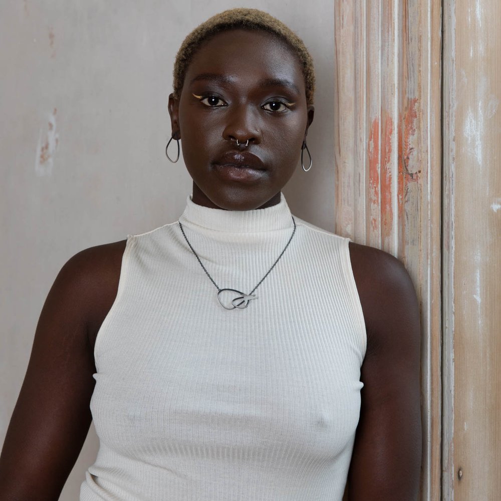 A person with short, blonde hair and hoop earrings stands against a weathered wooden door frame. They are wearing a white sleeveless turtleneck and a necklace with an oxidised silver circular pendant featuring bird designs. They have a septum piercing and are gazing directly at the camera, flaunting a Two Tone Silver Wing Pendant Necklace with Birds by Leoma Drew.