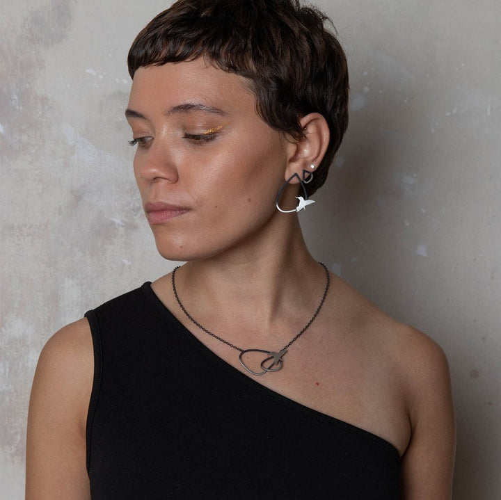 A person with short hair, wearing gold eyeliner and a black one-shoulder top, looks to the side. They are adorned with geometric earrings showcasing bird designs, and a matching Two Tone Silver Wing Pendant Necklace with Birds by Leoma Drew featuring similar geometric shapes. The accessories, crafted from oxidized silver, stand out against the plain and neutral background.