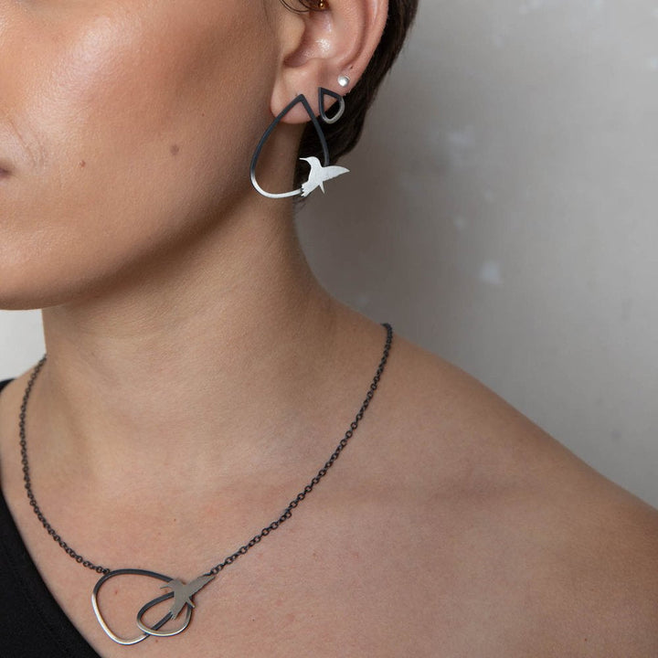 Close-up of a person wearing a matching jewelry set, featuring Leoma Drew's Two Tone Silver Wing Pendant Necklace with Birds and earrings shaped in abstract, delicate forms resembling birds in flight. The large earrings have a small bird detail, while the necklace showcases a similar bird motif.