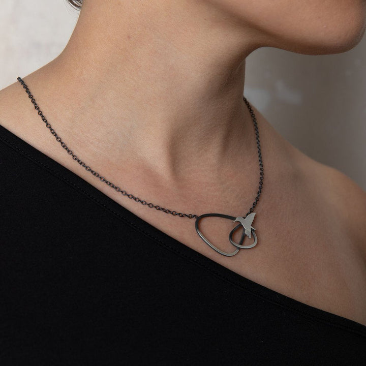 Close-up of a person wearing a one-shoulder black top and a delicate necklace. The Leoma Drew Two Tone Silver Wing Pendant Necklace with Birds features an abstract design with interlocking loops and a small star-shaped charm. The person's skin is softly illuminated, highlighting the jewelry's intricate details.