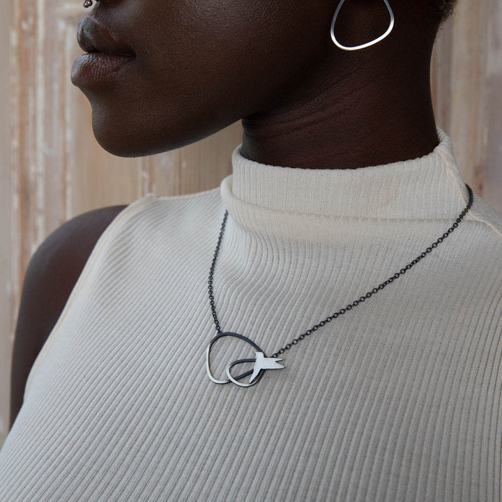 A person wearing a sleeveless, ribbed, white top displays a **Two Tone Silver Wing Pendant Necklace with Birds by Leoma Drew** on a black chain. They are also wearing a geometric, tear-shaped earring. The background features a textured, light-colored wall.