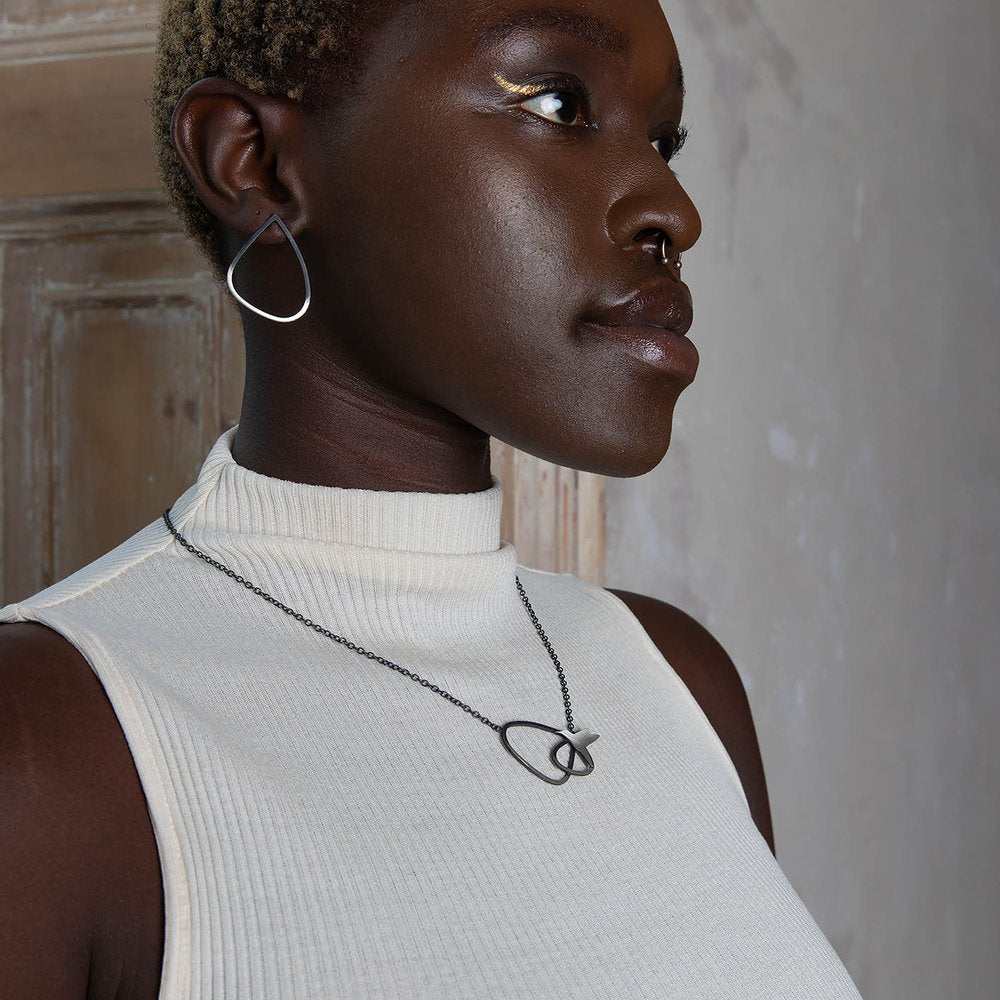 A person with short hair and dark skin wears a white sleeveless turtleneck top, a teardrop-shaped earring, and a Leoma Drew Two Tone Silver Wing Pendant Necklace with Birds. They are standing against a distressed wooden door background, looking away from the camera.