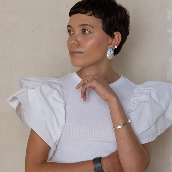 A person with short dark hair, wearing a white shirt with ruffled sleeves, looks to the side thoughtfully. They are accessorized with a Silver Kimana Cuff Bangle from Leoma Drew, a dark wristband, a ring, and large geometric-shaped earrings. The background is a plain, light-colored wall.