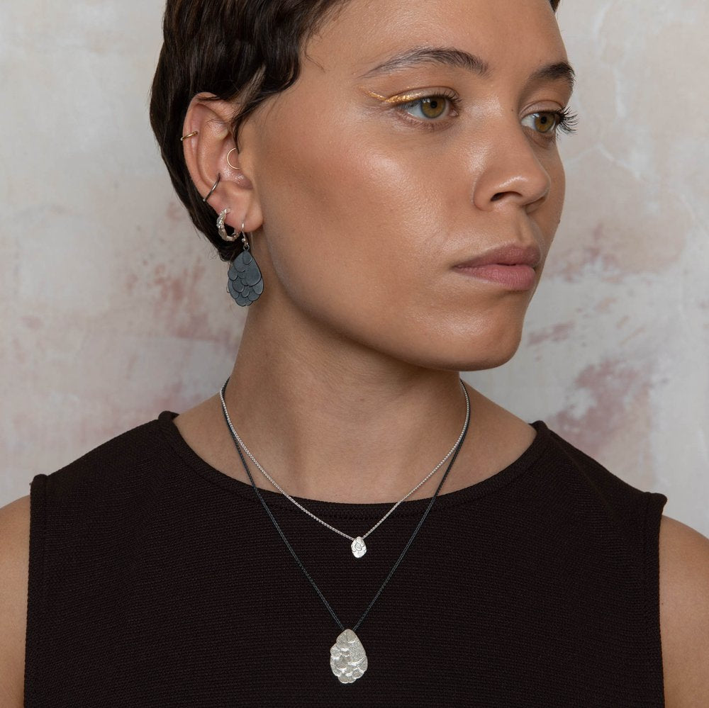 A person with short hair is wearing Leoma Drew Freeform Layered Wing Hoop Earrings in Frosted Silver and earrings with unique, textured pendants. Dressed in a black sleeveless top, they sport yellow eyeliner against a light, marble-like background.