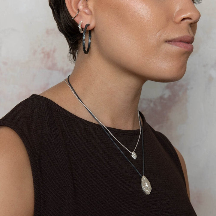 Close-up of a person from the neck up, wearing a dark sleeveless top. They have short hair and are adorned with two necklaces, one with a small round pendant and the other with a larger, irregular-shaped one. They are also wearing Leoma Drew Freeform Layered Wing Hoop Earrings in Frosted Silver with dangling elements.