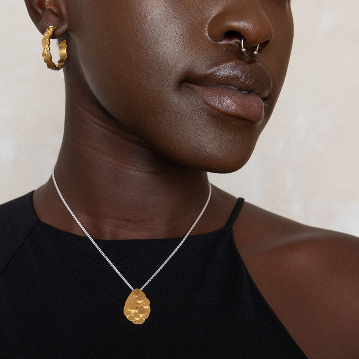 A person wearing a dark sleeveless top is shown from the chin down. They have a gold hoop earring, a nose ring, and a Kimana Necklace in Gold by Leoma Drew with a unique, textured design in gold vermeil. The background is neutral and out of focus.