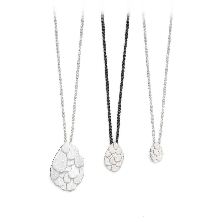 Three necklaces with pendants resembling overlapping scales come in three sizes. The left Leoma Drew Kimana Necklace in Frosted Silver features a large oval-shaped pendant on a silver chain. The middle pendant is medium-sized on a black chain, while the right has a small oval pendant on a frosted silver chain. All boast a metallic finish.