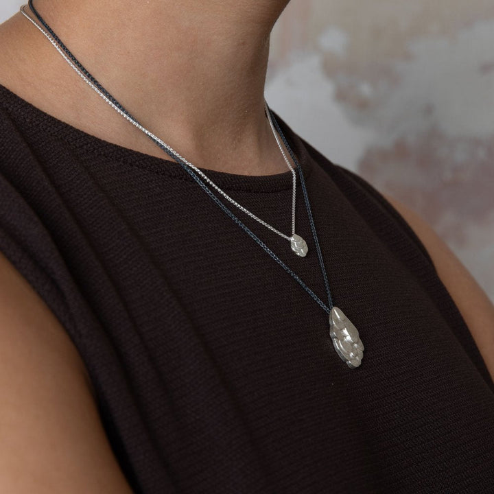 Close-up of a person wearing a black sleeveless top and two Leoma Drew Kimana Necklaces in Frosted Silver, both featuring small, translucent pendants. The background is blurred with soft, muted colors. The focus is on the frosted silver necklaces and the neckline of the top.