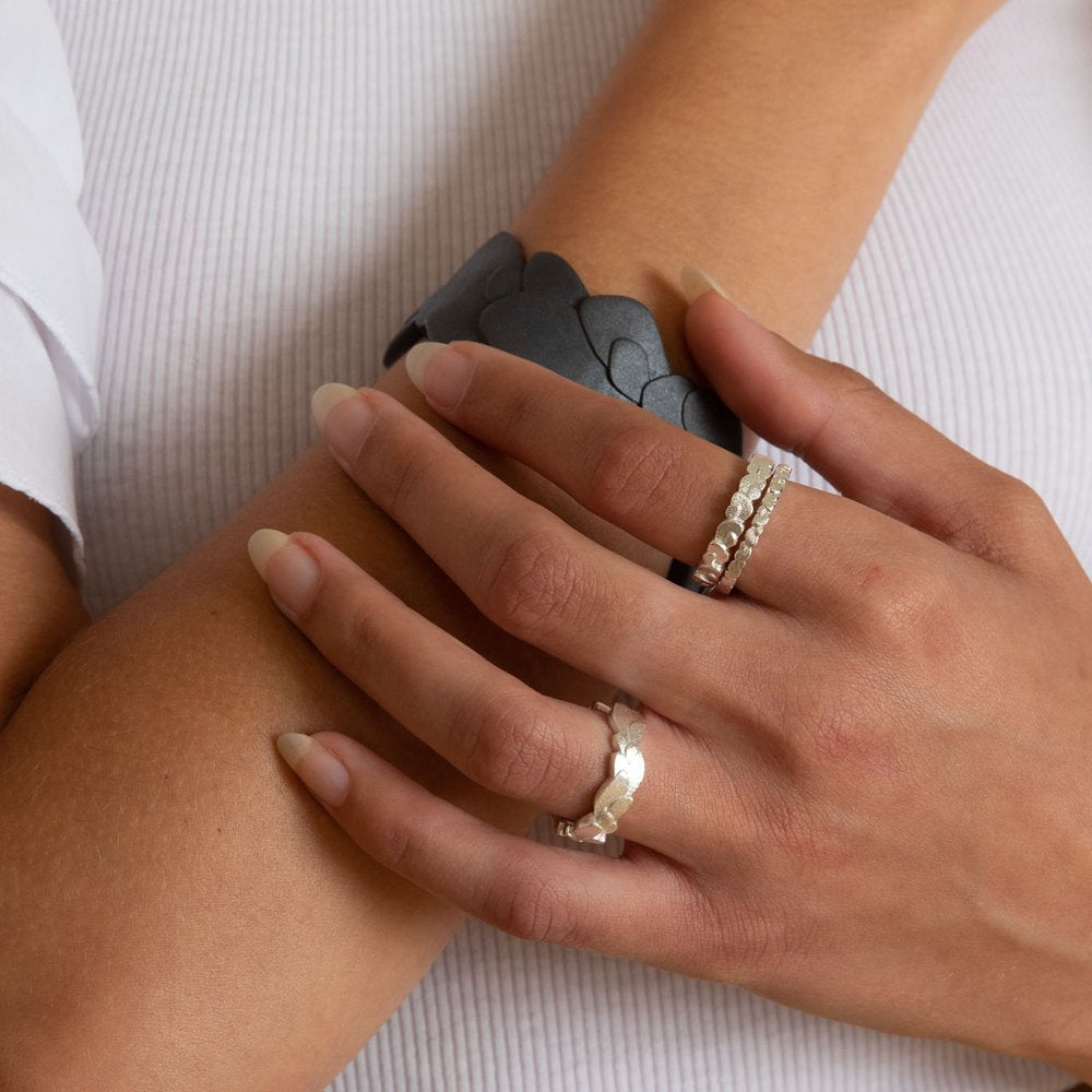 A close-up image of two hands resting on a white ribbed fabric surface. One hand wears three frosted silver rings with different designs, including the **Kimana Silver Double Wing Ring** from **Leoma Drew**, and the other hand has natural, unpolished nails. An arm in the background wears a black, leaf-patterned bracelet from the Kimana collection.