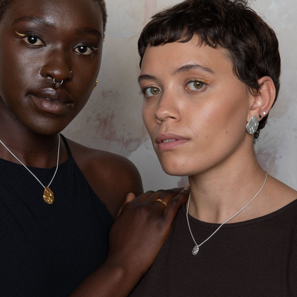 Two people standing closely together. One has dark skin, a nose ring, and wears a Leoma Drew Kimana Necklace in Frosted Silver and black top, touching the shoulder of the other, who has light skin, short hair, and wears a frosted silver pendant necklace and brown top. Both look towards the camera.