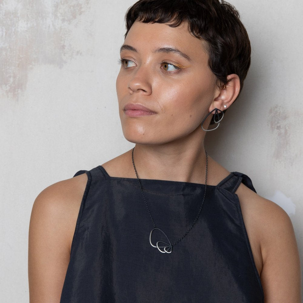 A person with short dark hair is wearing a sleeveless dark gray dress, matching geometric earrings, and a necklace featuring the Leoma Drew Two Tone Triple Wing Pendant. They are looking to the right against a light textured background.