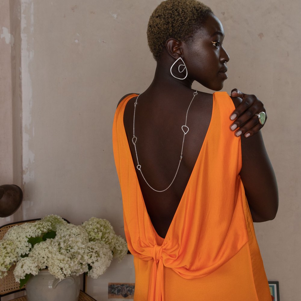 A person with short, light hair is seen from the back wearing an orange, backless dress adorned with a long chain necklace that drapes down their back. They are also wearing Leoma Drew's XL Wing Studs with Medium Wing Insert Frosted Silver and a ring. White flowers in a vase are visible in the background.