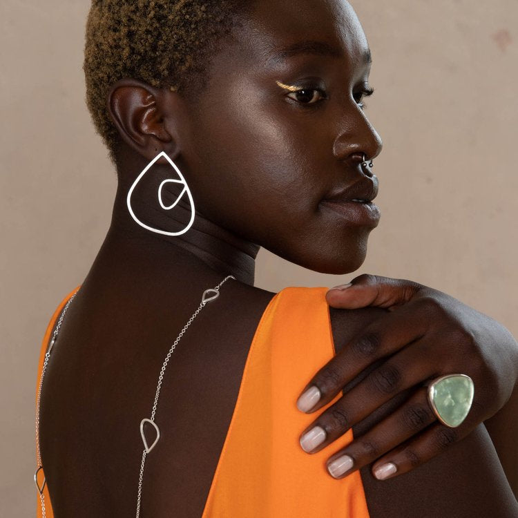 A person with short hair is showcasing jewelry while wearing an orange sleeveless top. The handmade earrings, identified as Leoma Drew’s XL Wing Studs with Medium Wing Insert in Frosted Silver, include an abstract white drop design. These are complemented by a delicate back necklace and a large green ring. The background is neutral, highlighting the vibrant attire and accessories.