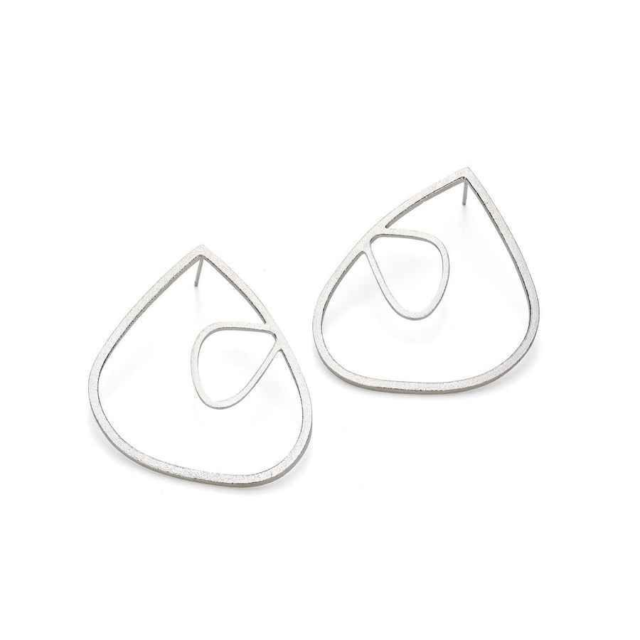 The XL Wing Studs with Medium Wing Insert Frosted Silver by Leoma Drew are handmade, silver-toned earrings featuring a modern, abstract design. Each earring showcases a large teardrop-shaped outer frame that encloses a smaller, irregular loop within it. The minimalist style emphasizes clean lines and contemporary aesthetics, making them perfect for those who appreciate sterling silver earrings.