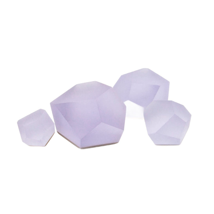 Four faceted, lavender-colored geometric shapes are arranged on a white background, resembling ring tops. The shapes vary in size, with the largest on the right and the smallest on the left. The smooth surfaces have a subtle gradient, giving them a slightly translucent appearance. These are Fruit Bijoux Ring Top VU Crystals Lilac Purple by Fruit Bijoux.