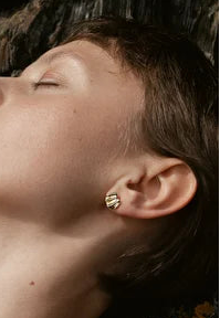 A person with short brown hair and red lipstick, eyes closed and head tilted back, wears Inner Island's A Little Joy Earrings. The background is rocky, adorned with orange and yellow lichen.