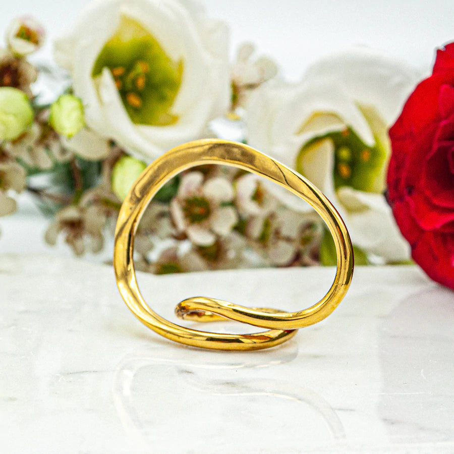 An Infinity Ring Gold, crafted by Lotta et Djossou, is displayed on a white marble surface, with delicate white and red flowers blurred in the background. The ring features a simple, elegant design with a smooth, twisting loop.