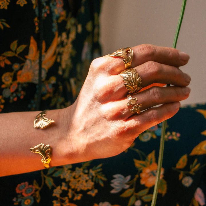 A hand adorned with gold jewelry featuring leaf designs, including an intricate adjustable ring and a bracelet, set against a background of a dark floral patterned garment. The lighting casts a warm glow on the hand and the Lotta et Djossou Coco Feather Ring Gold.