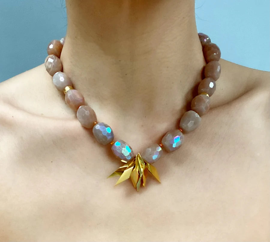 A person wears the Aria V Lotus Necklace, featuring large faceted sunstone beads and a central gold pendant with layered petals. Handmade in Dublin, this standout piece contrasts beautifully against a plain light blue background.