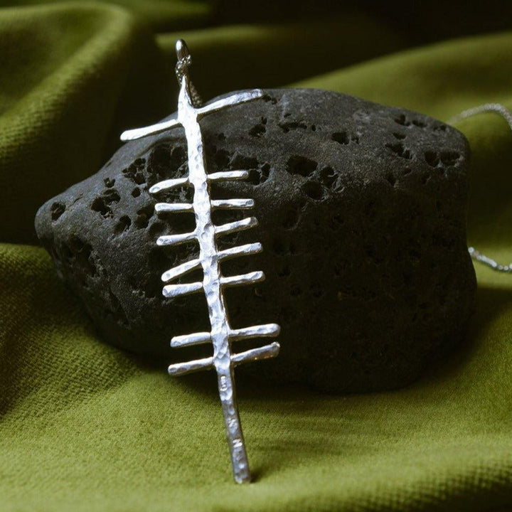 A Love (Grá) Large Rustic Pendant Silver from Ogham Treasure lays against a rough, textured black volcanic rock, placed on a green fabric background. Inscribed with "Grá," the pendant has a hammered finish, giving it a rustic look.